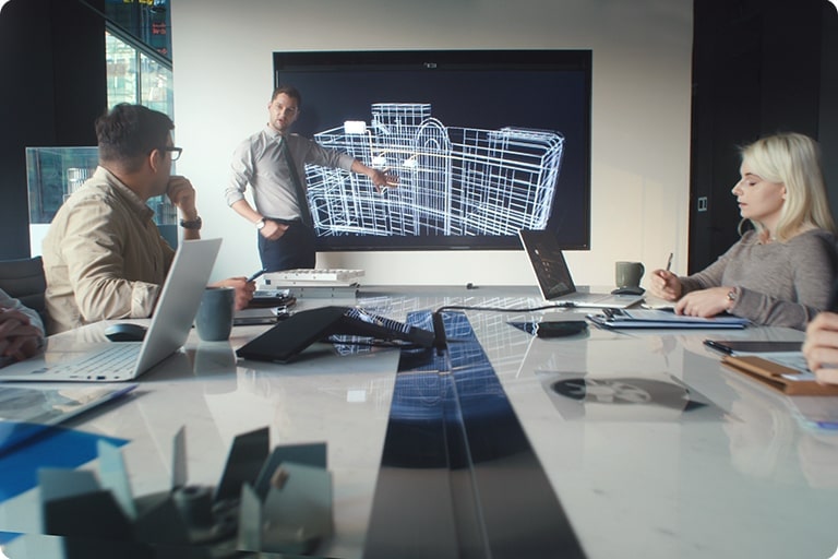 Em uma sala de conferências, quatro pessoas sentam-se enquanto um homem fica no centro, apontando para uma tela que exibe a planta de um edifício.
