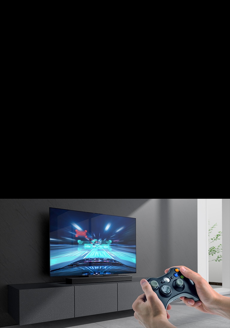 La barra de sonido se coloca en el gabinete y la escena del juego de carreras se muestra en el televisor conectado a la barra de sonido. Una consola de juegos está en la parte inferior derecha de la imagen sostenida con dos manos.