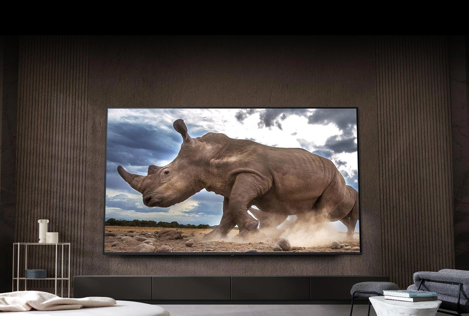Auf einem Ultra Big LG-Fernseher, der an der braunen Wand eines Wohnzimmers montiert ist, umgeben von cremefarbenen modularen Möbeln, wird ein Nashorn in einer Safari-Umgebung gezeigt.