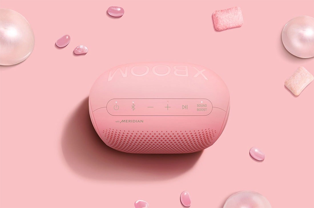 LG XBOOMGo PL2P Bluetooth Speaker, Kaugummis, Jellybeans und ein LG XBOOM Go PL2P sind auf einem rosa Hintergrund platziert., PL2P