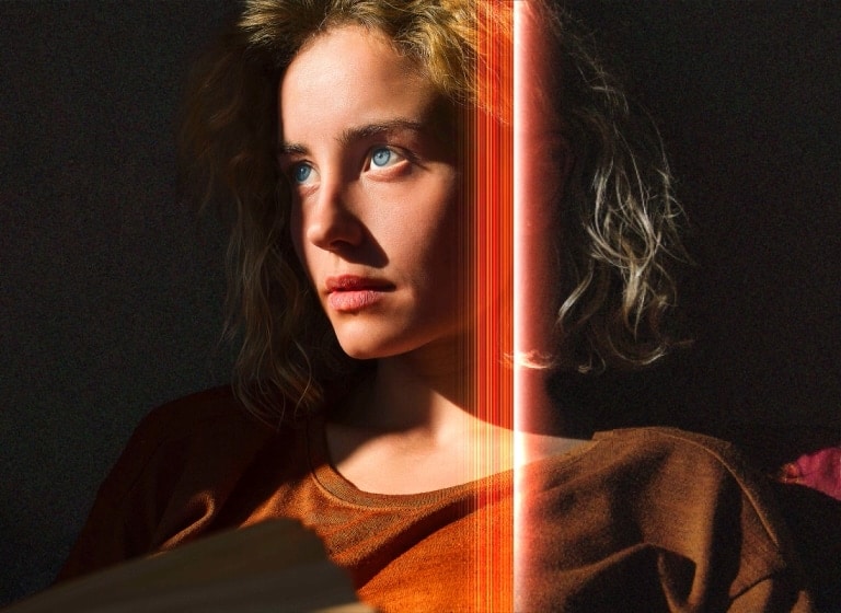 Eine Frau mit strahlend blauen Augen und einem orangefarbenen Oberteil in einem dunklen Raum. Rote Linien, die AI-Verfeinerungen darstellen, bedecken einen Teil ihres Gesichts, das hell und detailliert ist, während der Rest des Bildes stumpf aussieht. 
