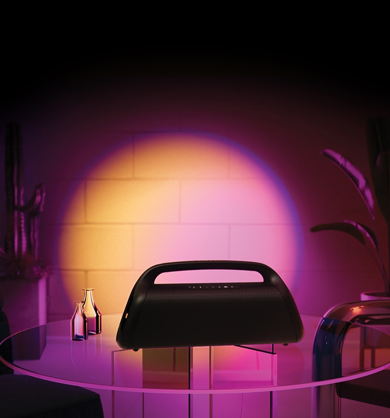 Uma imagem da LG XBOOM Go XG9. Ela está colocada numa mesa de vidro em casa, projetando iluminação de palco em degradê de rosa e laranja.
