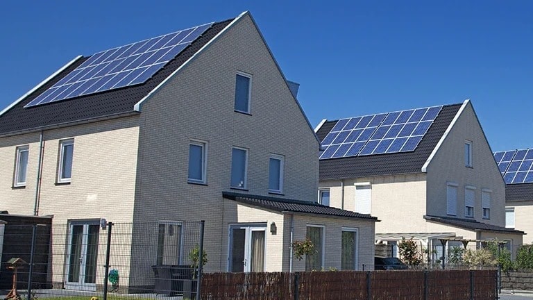 A vista frontal das casas em tom marfim é exibida lado a lado com o telhado preto coberto por painéis solares.