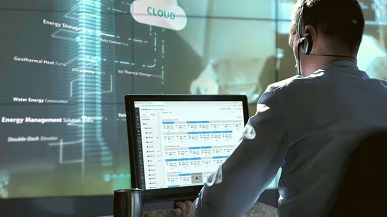 Um homem com fones de ouvido olha para um monitor que exibe dados variados tendo como pano de fundo uma tela conectada a um servidor em nuvem.