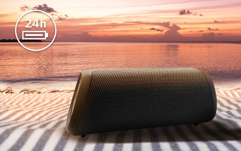 A caixa de som está colocada sobre uma toalha de praia. Na frente dela, vê-se uma praia ao pôr do sol para ilustrar que a caixa de som pode ficar ligada por até 24 horas.