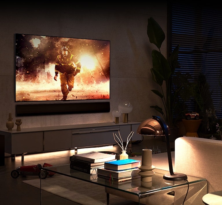 Numa sala espaçosa e confortável, TV e soundbar estão montadas na parede Na tela da TV, vê-se um bombeiro pulando para fora de um prédio em chamas.