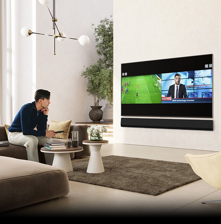 Un hombre sentado en un lujoso y espacioso sofá de la sala de estar, mirando la televisión. La pantalla del televisor muestra una pantalla dividida de un partido de fútbol y un programa de noticias.