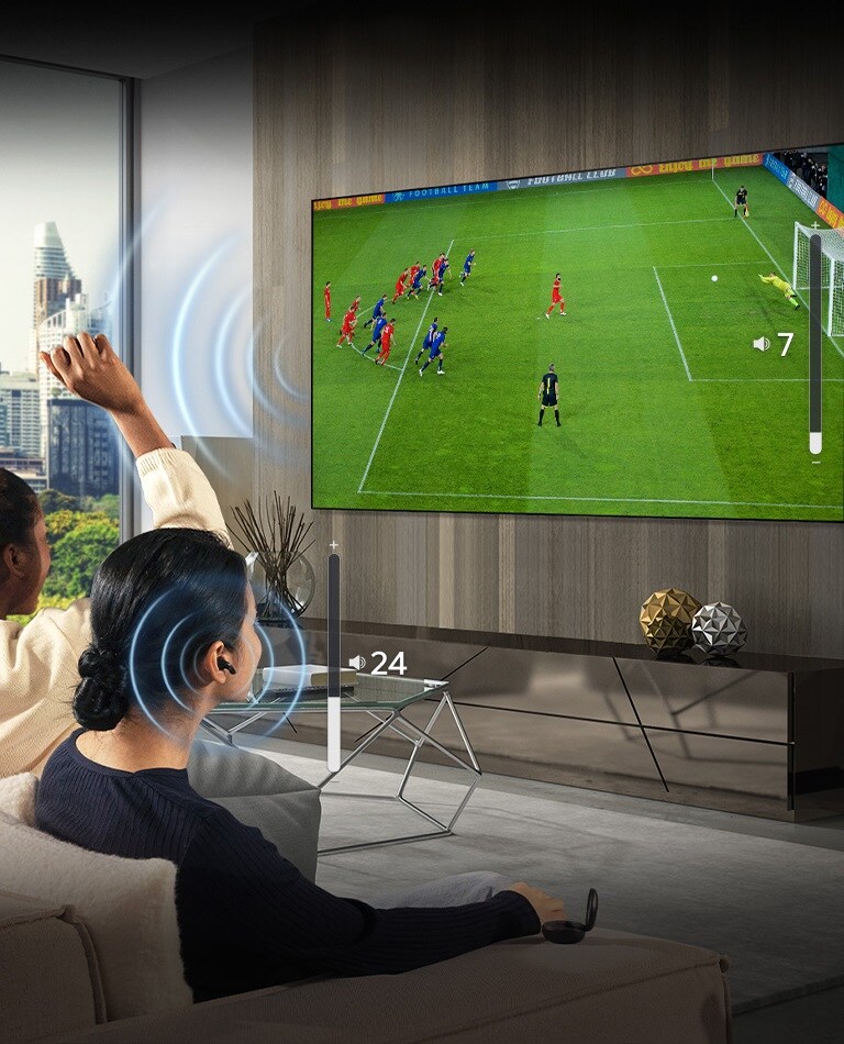 Un grupo de personas están sentadas en un sofá viendo un partido de futbol en la televisión. La mujer situada en el extremo derecho tiene puestos unos auriculares y los utiliza con un volumen distinto al de la televisión, lo que indica que está utilizando ambos al mismo tiempo.