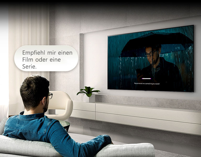Ein Mann sitzt auf einem Sofa und blickt auf einen Fernseher. Über seinem Kopf befindet sich eine Sprachblase mit den Worten „Empfiehl mir einen Film oder eine Serie.“ Die Szene auf dem Bildschirm zeigt einen Mann mit Regenschirm, außerdem ist die Benutzeroberfläche der Spracherkennung zu sehen.