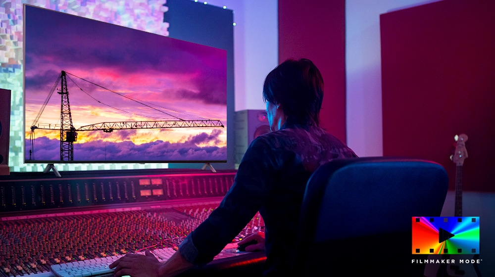 Un director de cine está mirando un gran monitor de TV, editando algo. La pantalla de TV muestra una grúa en el cielo púrpura. El logotipo de FILMMAKER Mode se coloca en la esquina inferior derecha.