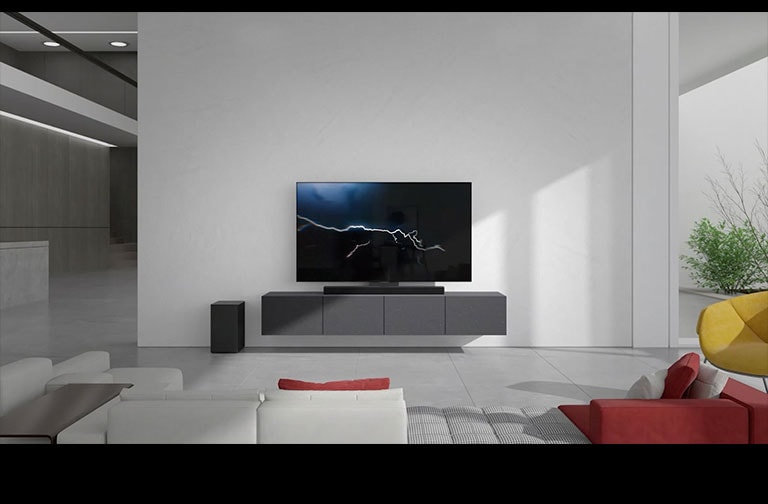 A barra de som está posicionada num armário cinzento com uma TV na sala de estar. Um subwoofer sem fios preto está posicionado no chão do lado esquerdo e a luz do sol entra pelo lado direito da imagem. Um sofá comprido branco e vermelho está virado para a TV e a barra de som.