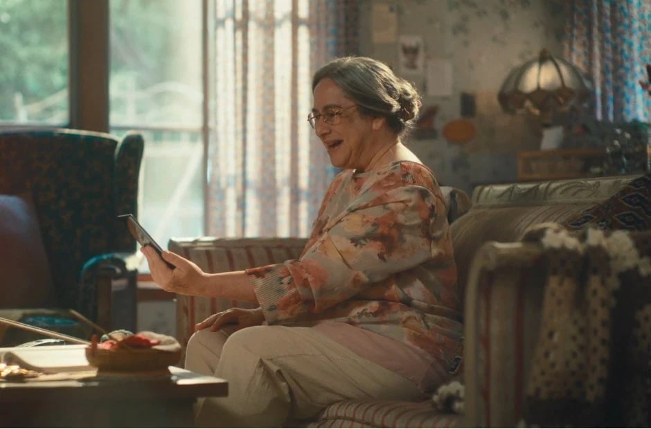 صورة تعرض امرأة مسنّة تجلس في غرفة معيشتها مبتسمة أثناء النظر إلى هاتف ذكي في يدها.