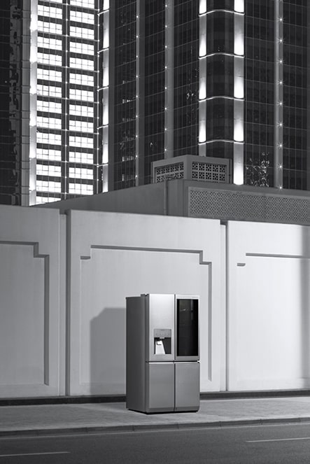 Der LG SIGNATURE Kühlschrank steht in der Nacht auf einer Straße und aus den Fenstern der Gebäude dringt Licht.