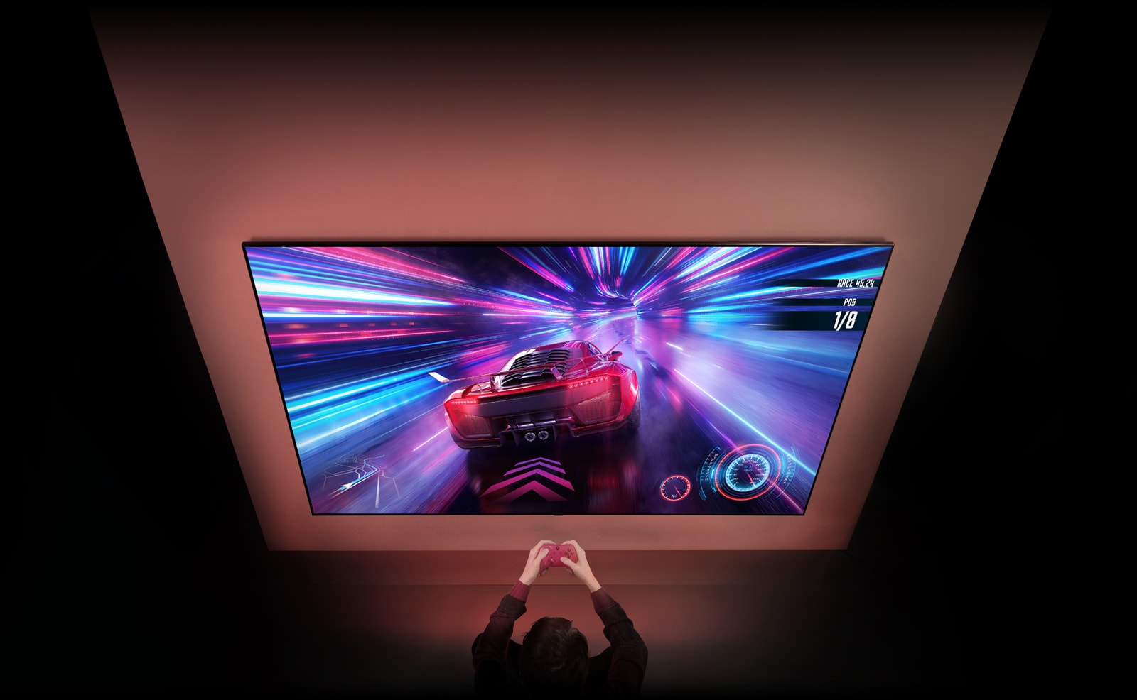Der er et stort tv på væggen, og på skærmen vises et bilspil, der er i gang. Foran tv'et står en spilcontroller i hænderne på en person, der er fokuseret på spillet.