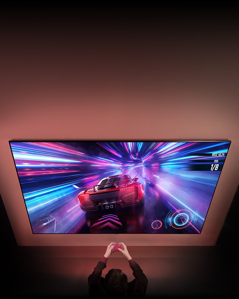 Der er et stort tv på væggen, og på skærmen vises et bilspil, der er i gang. Foran tv'et står en spilcontroller i hænderne på en person, der er fokuseret på spillet.