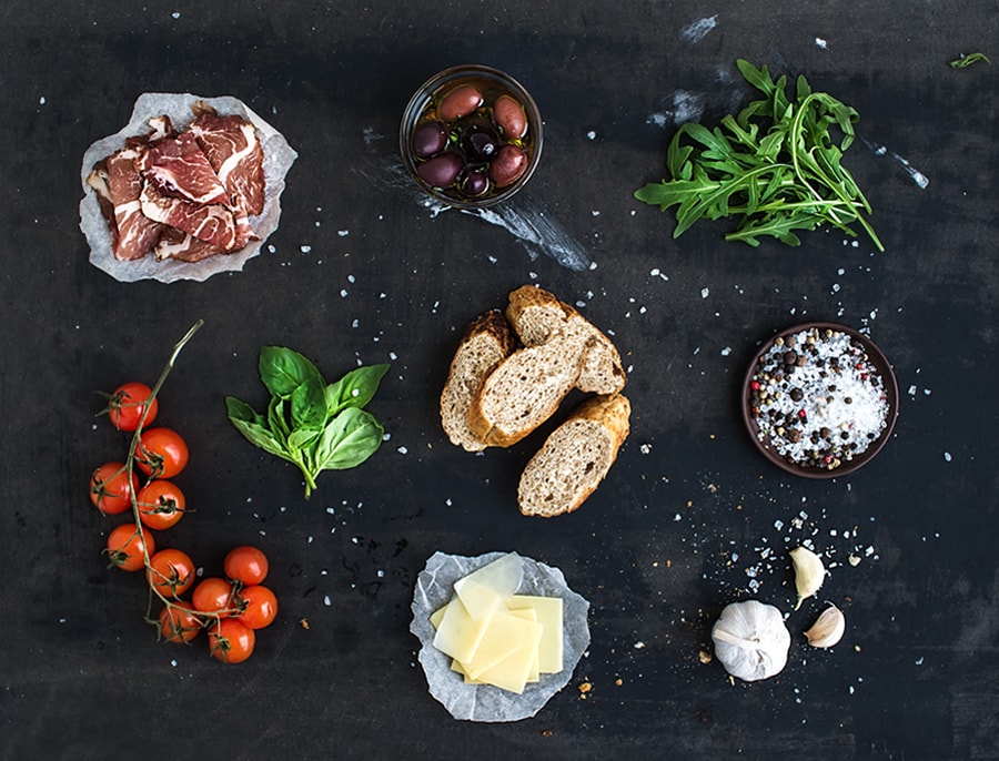 An array of fresh ingredients, including meat, olives, tomatoes, garlic, bread, herbs, and spices on a black surface