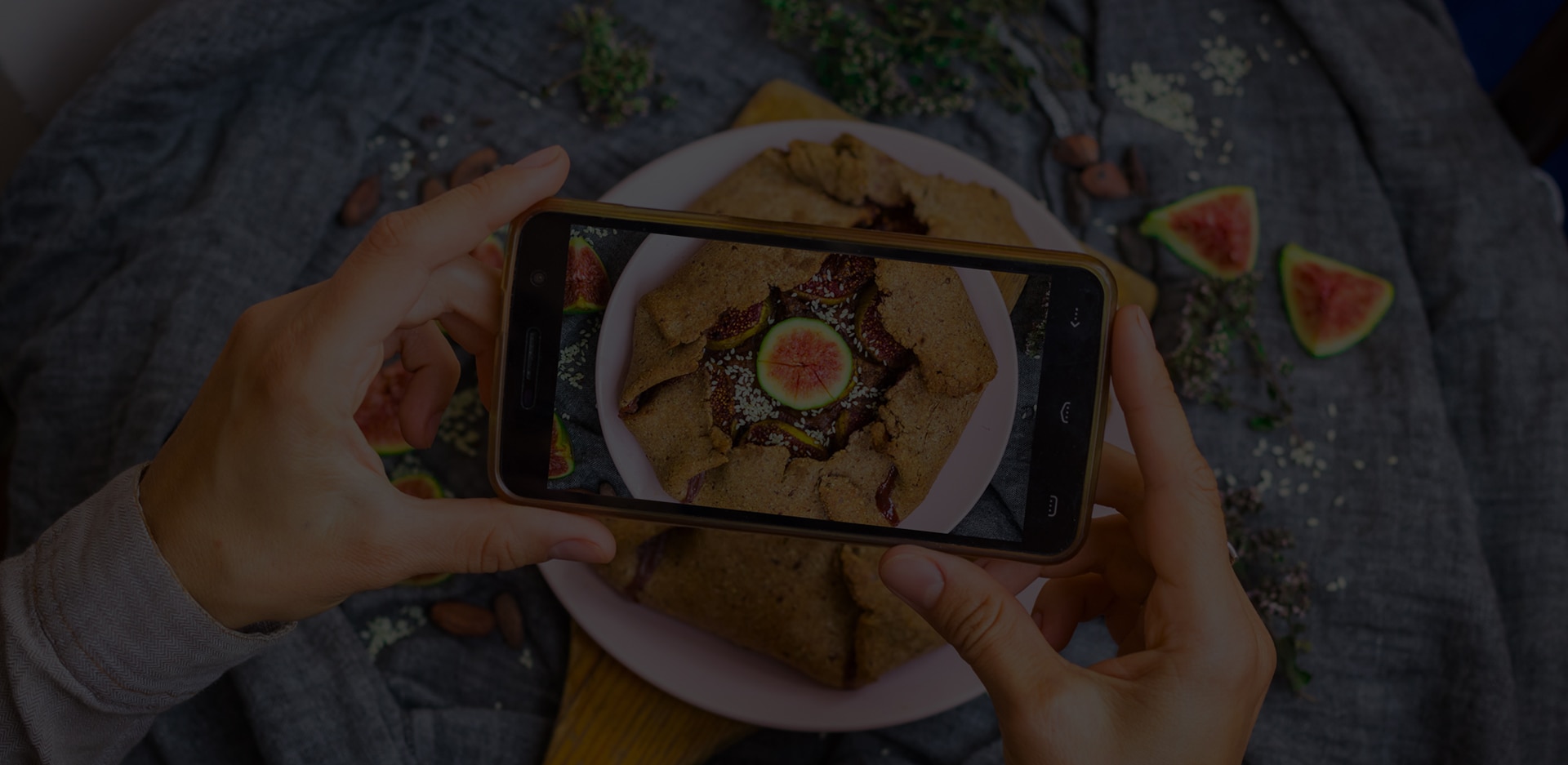 A pair of hands hold a smartphone over a fig pastry while snapping a picture. 'We are what we eat' is superimposed over it.