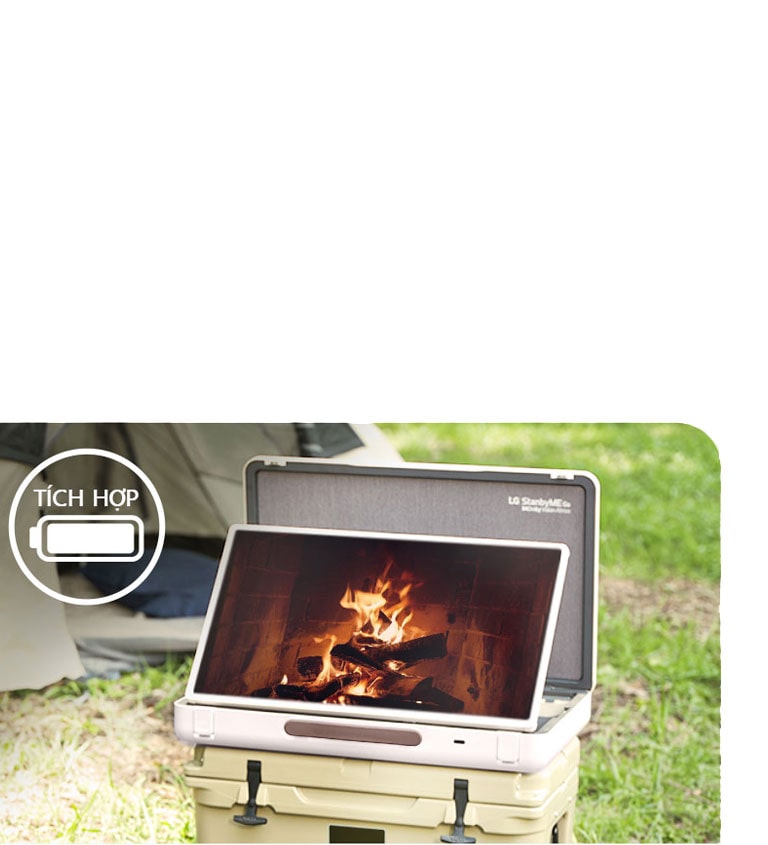 LG StanbyME Go được đặt trước một chiếc lều và màn hình hiển thị chủ đề thư giãn (lò sưởi). Ở góc trên cùng bên trái, biểu tượng pin tích hợp được hiển thị. 