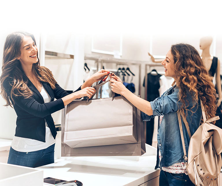 An Image of a sales clerk smiling and giving a customer shopping bags in a clothing store.