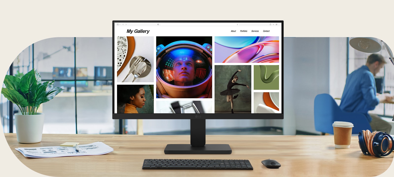 A modern desktop workspace featuring an LG monitor displaying a vibrant photo portfolio. The monitor is centered on a clean wooden desk with a keyboard, mouse, coffee cup, headphones, and potted plant. In the background, a man works near a large window in a contemporary office setting.