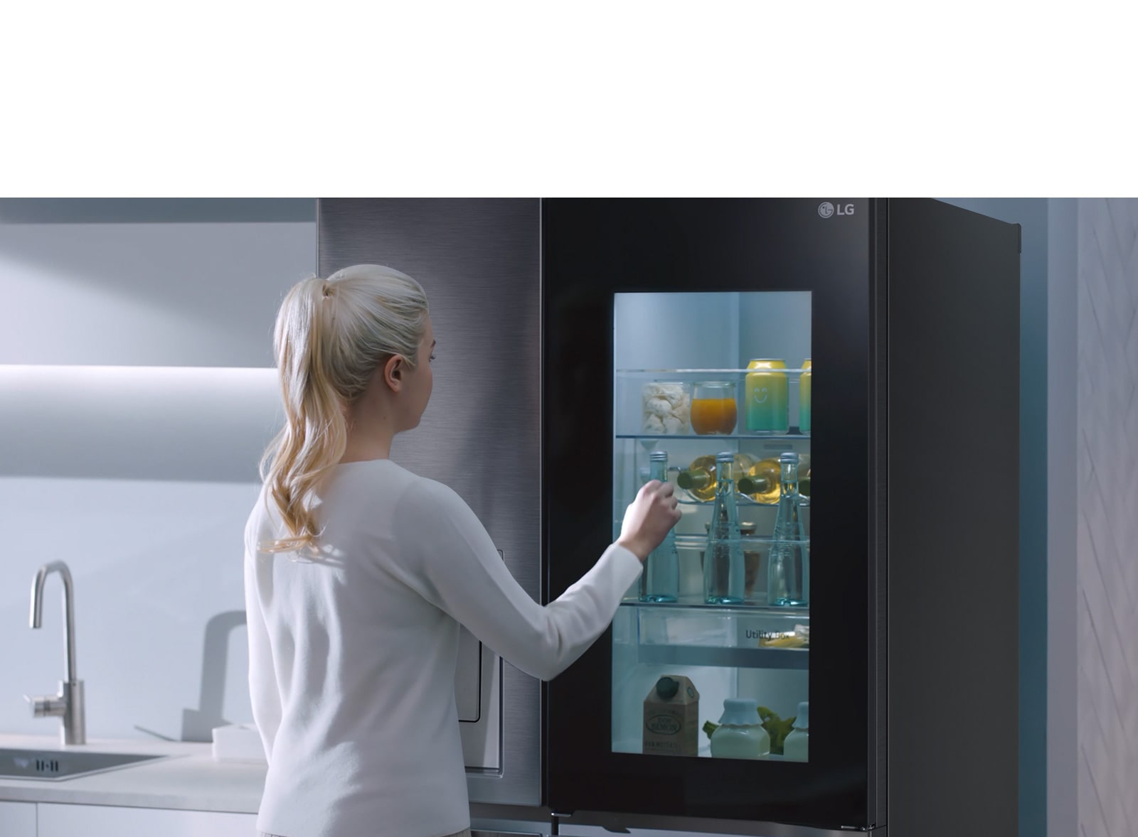 A video shows a woman approach her InstaView refrigerator and knock twice. The interior lights up and she can see the contents of her fridge without opening the door. The view zooms in to focus on the drinks in the door and then zooms out to see the woman from behind as she opens the door and grabs a drink.