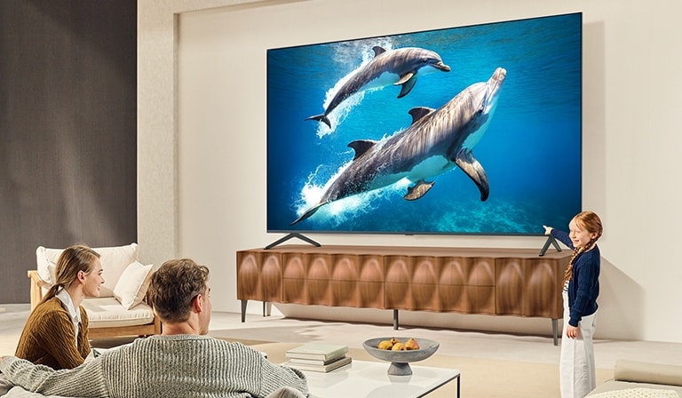 A family sits on a couch facing a LG QNED TV mounted on a wall above a LG Soundbar with a little girl pointing to a screen depicting two dolphins. 