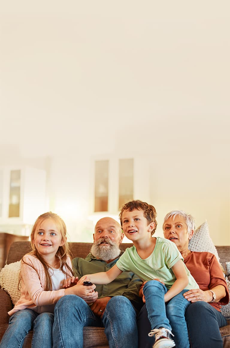 A family with children and their grandparents sits together on a sofa in a bright living room, holding a remote while watching TV. 