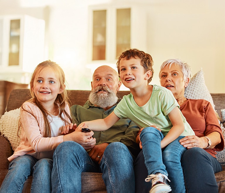 A family with children and their grandparents sits together on a sofa in a bright living room, holding a remote while watching TV.