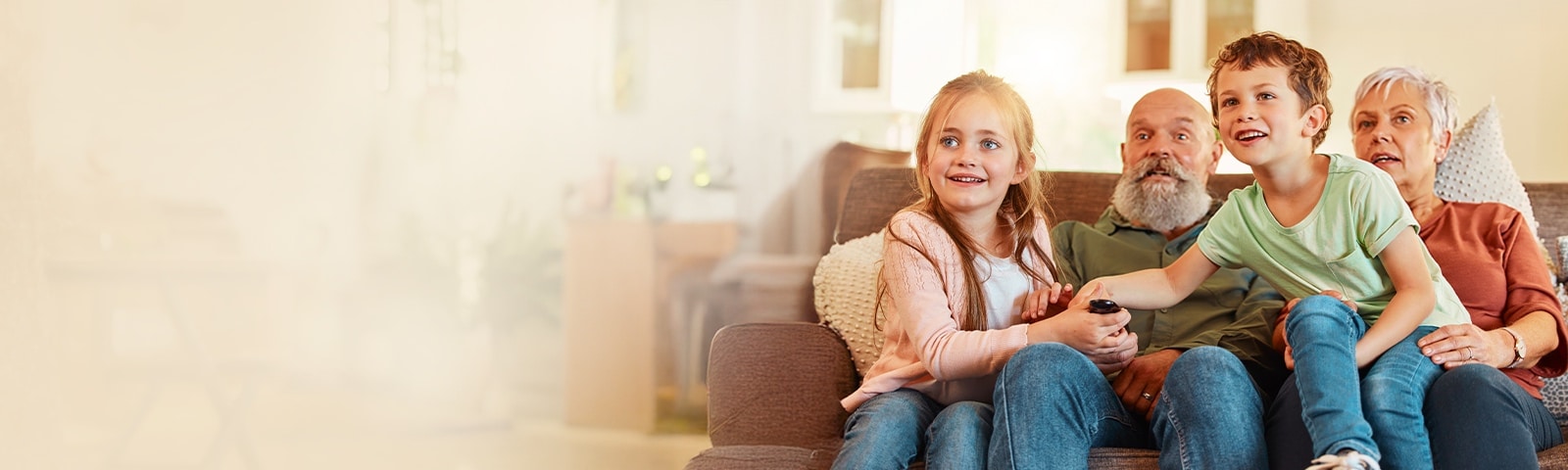A family with children and their grandparents sits together on a sofa in a bright living room, holding a remote while watching TV. 