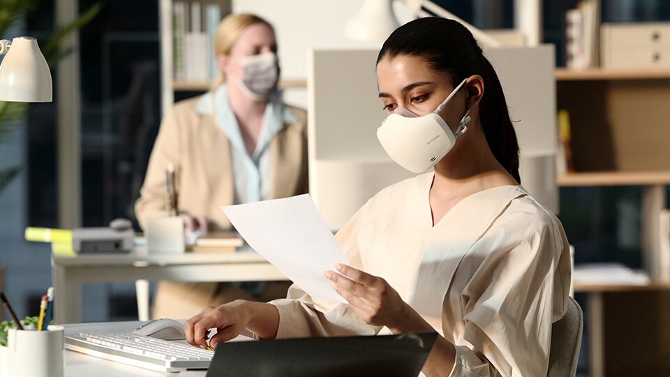 A woman is looking at documents in the office, with a white LG PuriCare™ Wearable Air Purifier on.