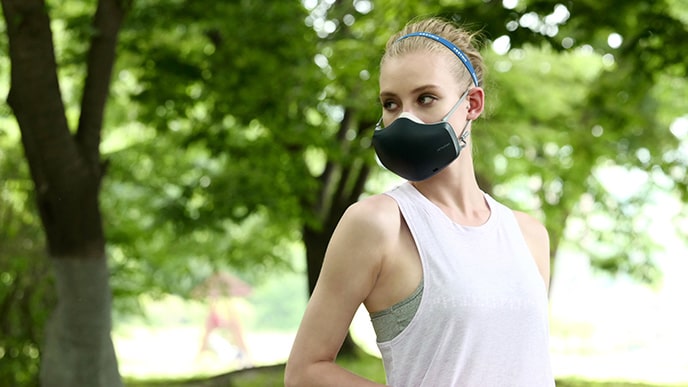 A woman who is wearing a black LG PuriCare™ Wearable Air Purifier seems to be resting after jogging in a park full of trees.