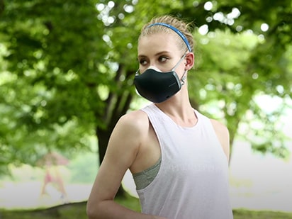 A woman who is wearing a black LG PuriCare™ Wearable Air Purifier seems to be resting after jogging in a park full of trees.