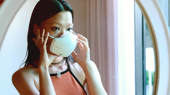 An Asian woman is wearing a white LG PuriCare™ Wearable Air Purifier while looking in the mirror.