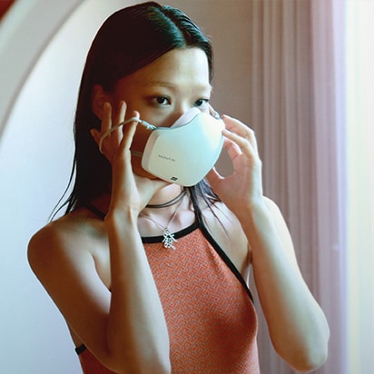 An Asian woman is wearing a white LG PuriCare™ Wearable Air Purifier while looking in the mirror.