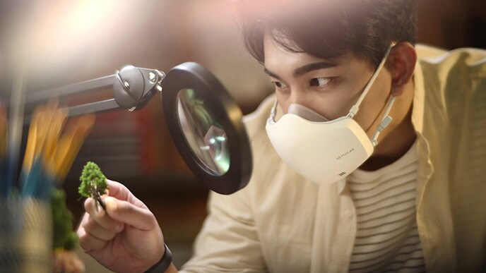 An Asian man is looking at something through a microscope, with a white LG PuriCare™ Wearable Air Purifier on.