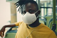 An African American man is sitting in a bus with a white LG PuriCare™ Wearable Air Purifier on while looking forward.