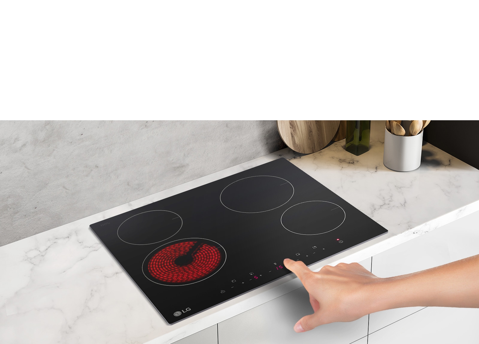 The STUDIO cooktop is seen from an angle up looking down. A hand reaches in to press a button. The left-front burner is red with heat.