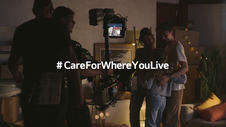 A film crew captures a family scene in a warmly lit living room, with the hashtag #CareForWhereYouLive displayed prominently in the center.