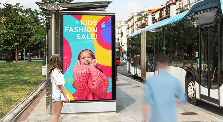 A large-sized display is installed at a bus stop, and a woman is looking at a vivid-quality advertisement on the screen.