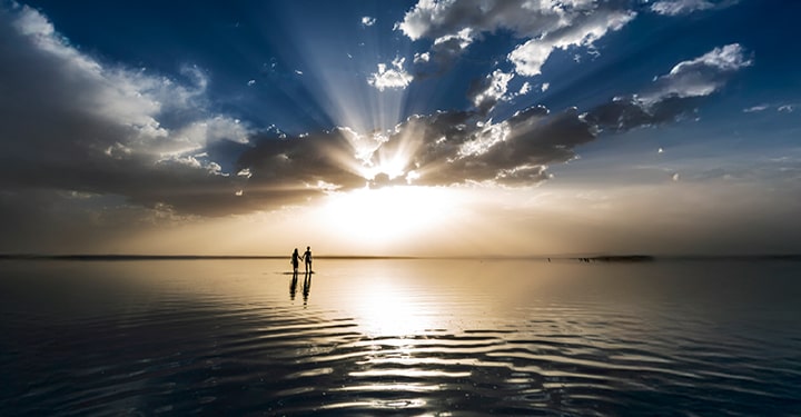 The sun shining above the horizon is partly hidden by clouds, casting rays of light across the dark sky, while its reflection on the water delicately contrasts with the surrounding darkness.