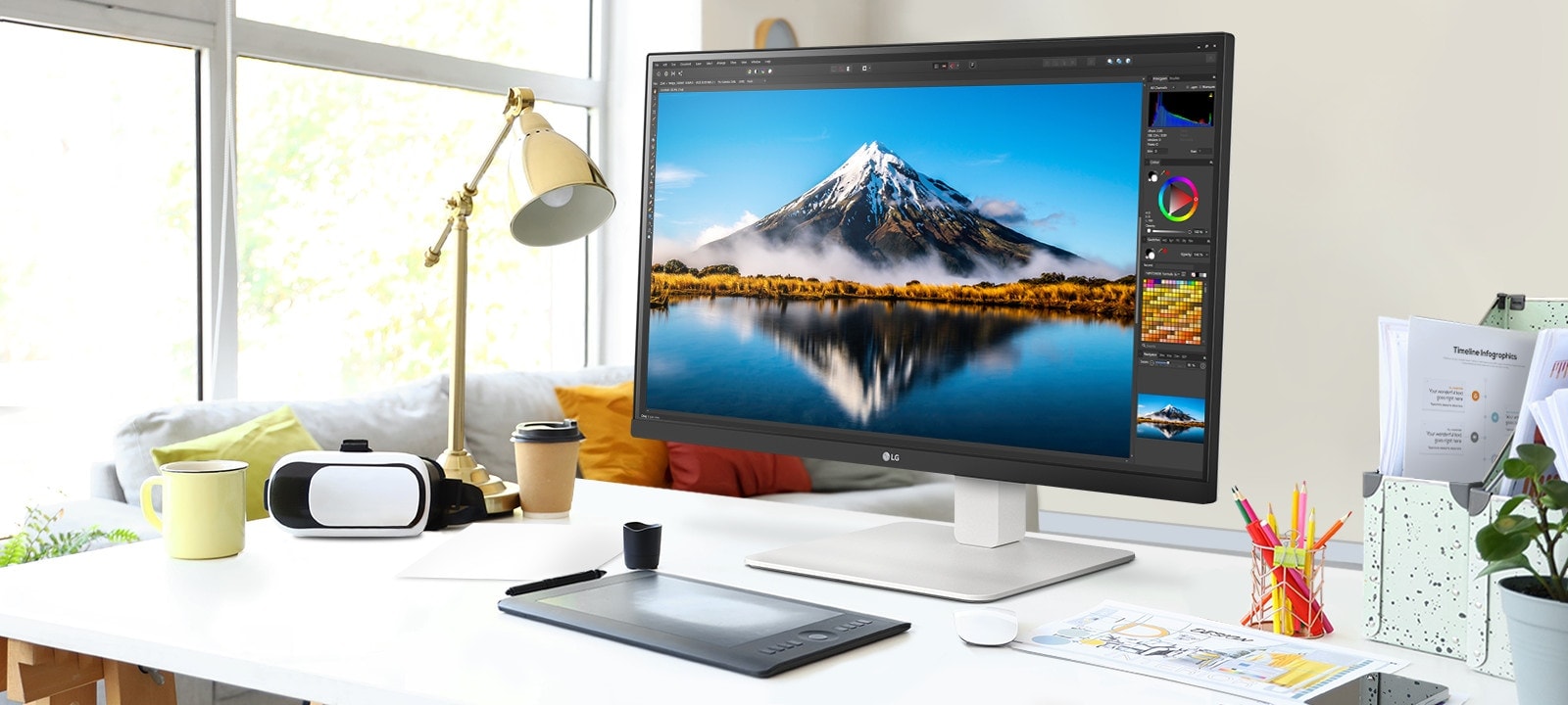 On the desk, there is an ultrafine monitor displaying Photoshop work. Next to the monitor, there is a desk lamp, a mouse, paper, and colored pencils.	