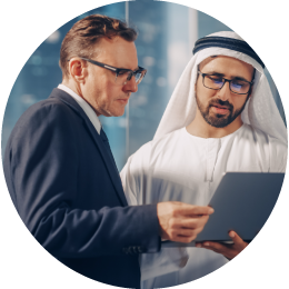 Two men in business attire, the left one in a suit and the right one in traditional Middle Eastern clothing, are discussing documents.