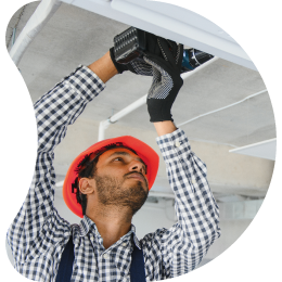 A technician wearing a red hard hat and safety gear, working on an overhead HVAC system.