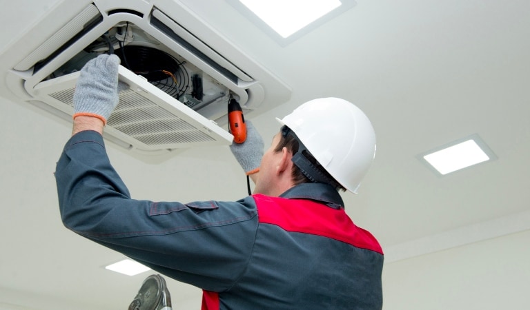 A technician wearing a hard hat and safety gear is working on an overhead HVAC unit.