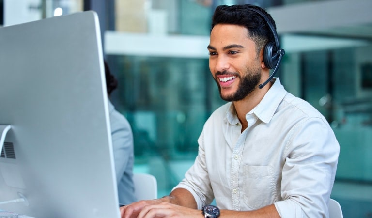 A smiling man wearing a headset is sitting in front of a computer, ready to assist with inquiries.