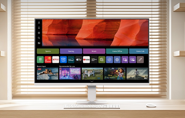 LG Smart Monitor on a clean wooden desk in a bright home office, displaying the webOS home screen with streaming apps and content tiles, paired with keyboard and mouse, bookshelf and window blinds in the background, highlighting smart entertainment and productivity use.