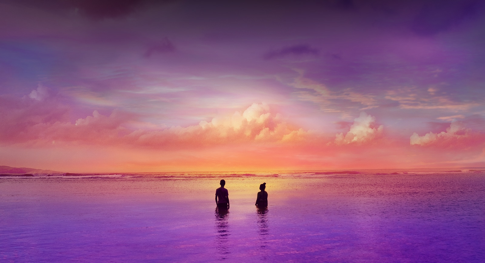 Two people are standing inside ocean – a slightly purple ocean. It’s a sunset time.