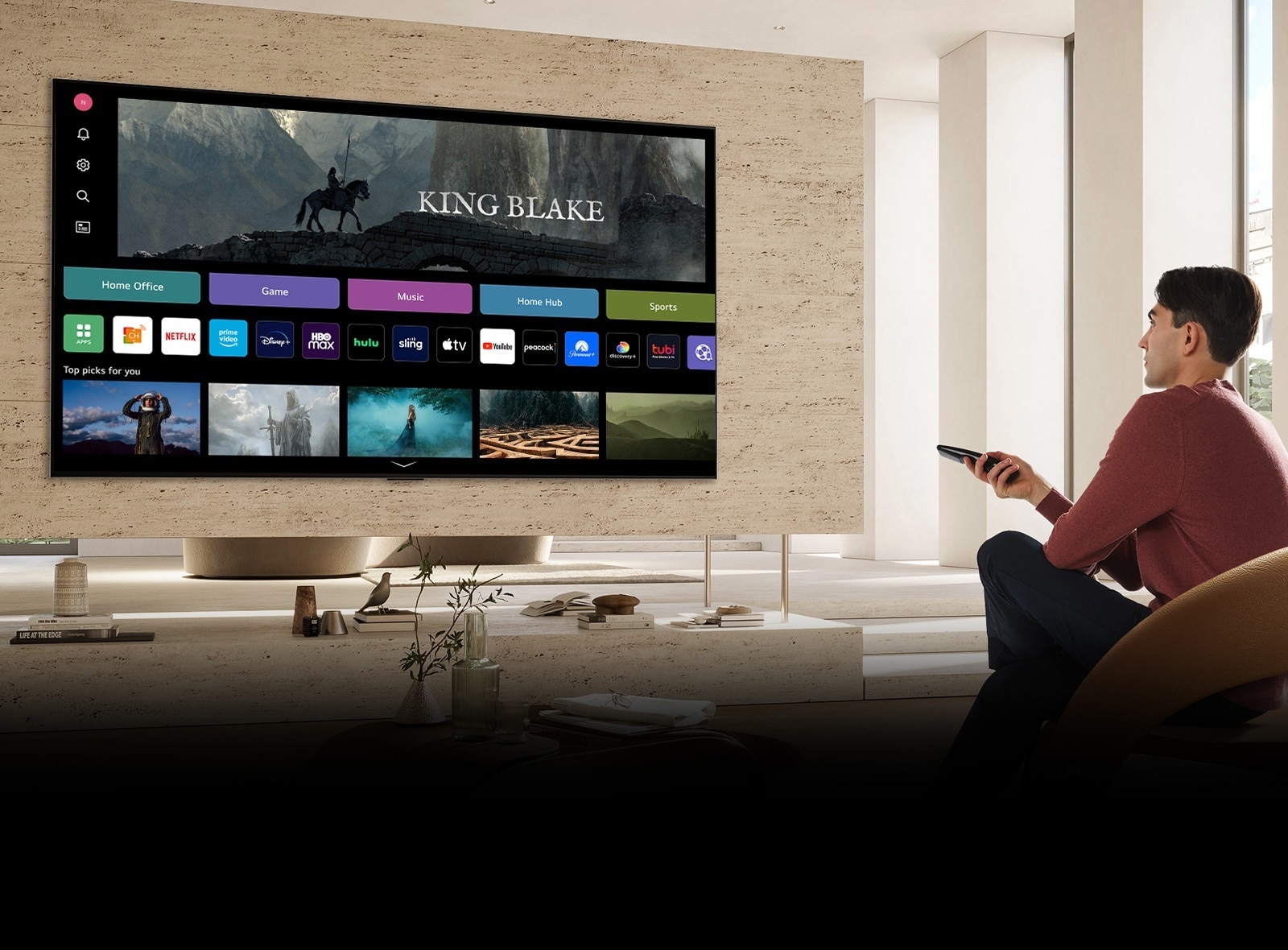 A man is holding a remote control in his right hand and looking at a large TV across from him. The TV screen shows the "All New Home" screen.