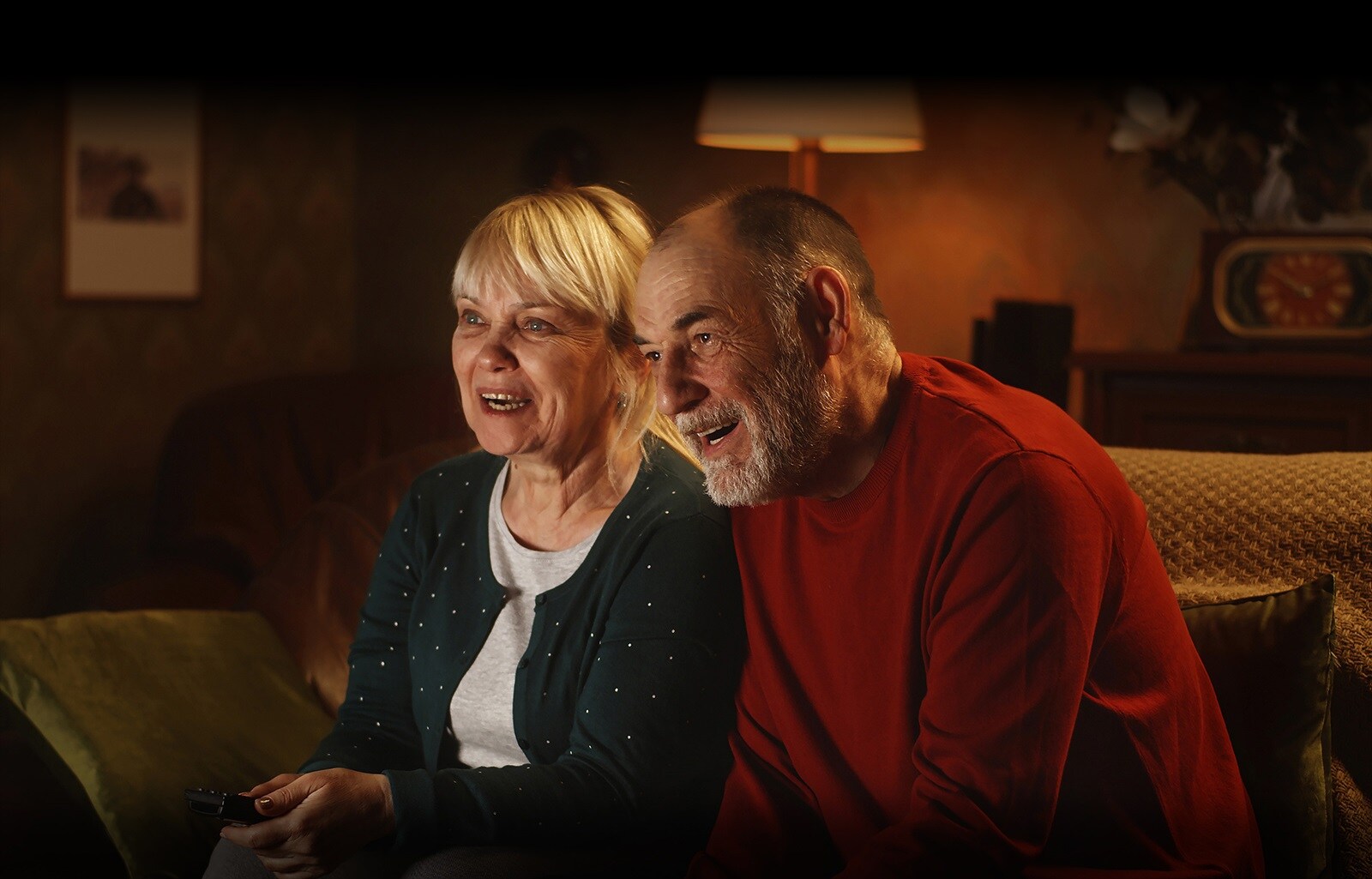 An elderly couple is sitting on a sofa, smiling and looking at the screen.