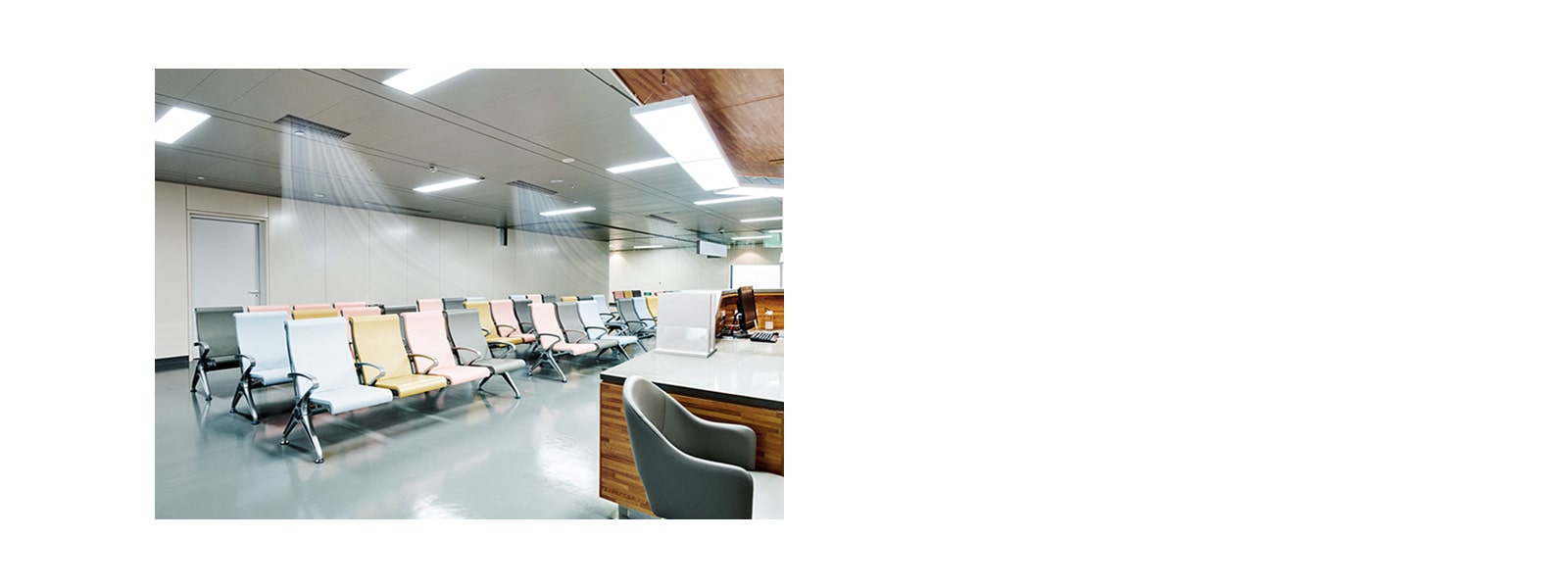 Three ceiling mounted air conditions create blue-colored air stream in a waiting room in hospital with a series of chairs and a reception desk to the right.